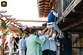 In Plovdiv, the groomsmen enthusiastically lift a man up to a wooden balcony as part of a spirited wedding tradition, infusing the celebration with an air of danger, excitement, and bold adventure against the backdrop of historic architecture.